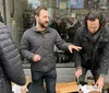 A group of people is engaged in a casual outdoor conversation one person gesturing expressively while another seems to be opening a box with urban surroundings and a reflective shop window in the background