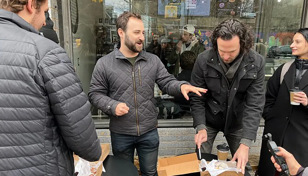 A group of people is engaged in a casual outdoor conversation one person gesturing expressively while another seems to be opening a box with urban surroundings and a reflective shop window in the background