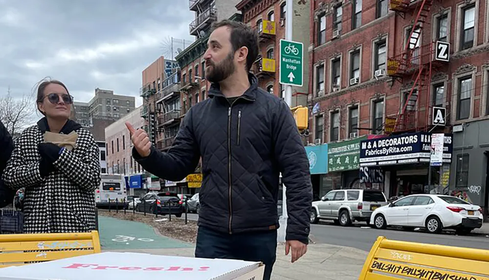 A man appears to be enthusiastically explaining something to a woman who is holding a sandwich while they stand on a city street with a ping pong table and various storefronts in the background