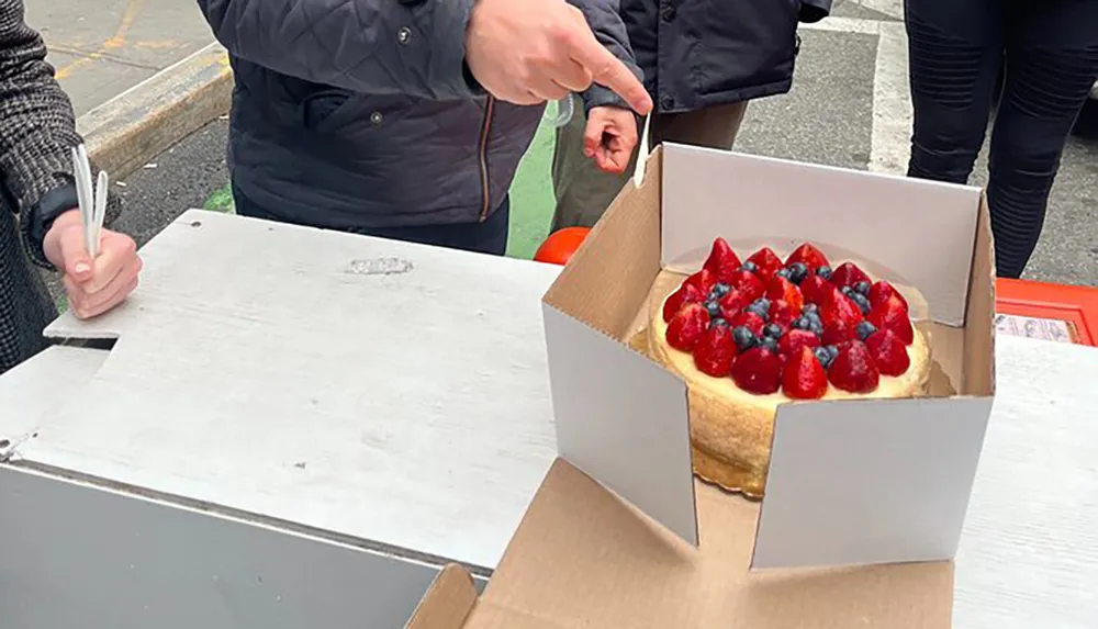 A person points to a freshly opened box containing a cheesecake topped with strawberries and blueberries as someone else watches