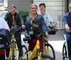 A group of smiling people are positioned on bicycles on an urban street wearing casual attire and conference lanyards