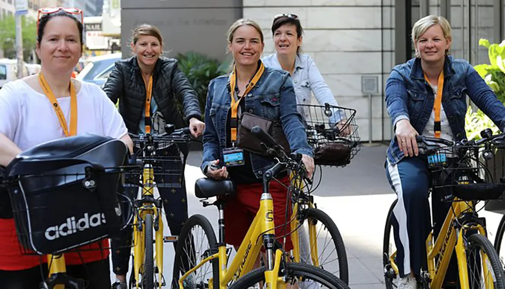 A group of smiling people are positioned on bicycles on an urban street wearing casual attire and conference lanyards