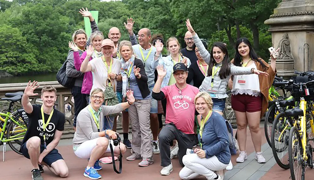 A diverse group of cheerful people with bicycles are posing for a photo with some waving or showing a thumbs-up possibly after a group cycling event