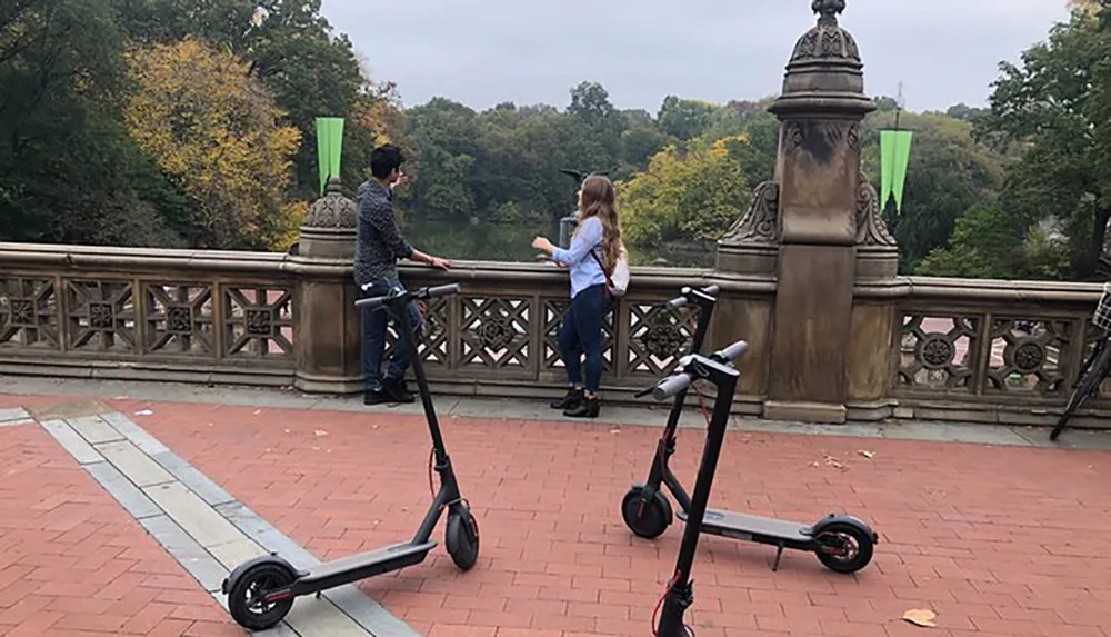 Two individuals are standing by a bridge railing and conversing with two electric scooters parked in the foreground against a backdrop of autumn foliage