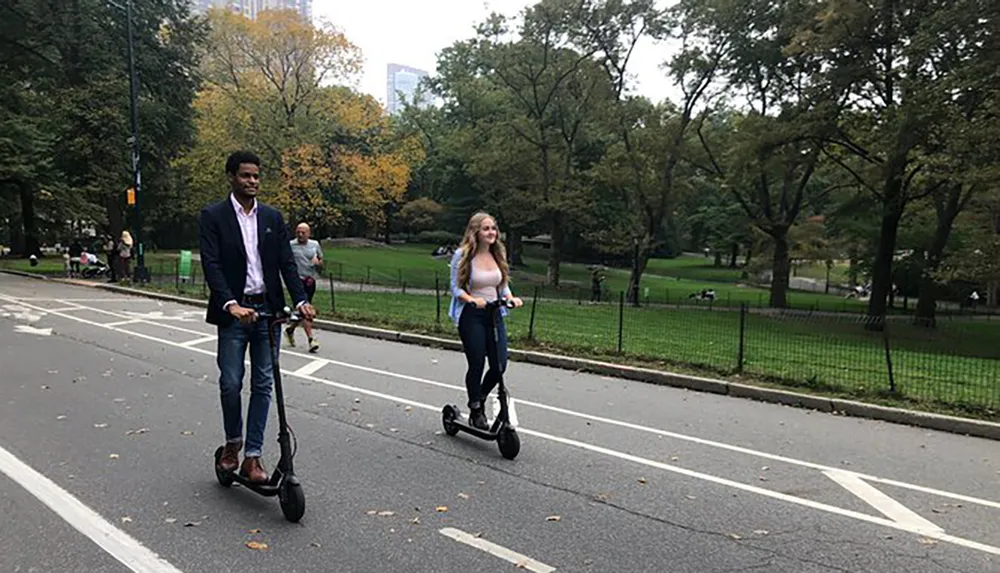 Two people are riding electric scooters on a park path surrounded by trees with the skyline in the background