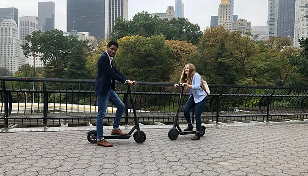 A man and a woman are smiling and riding electric scooters on a paved path with trees and city buildings in the background