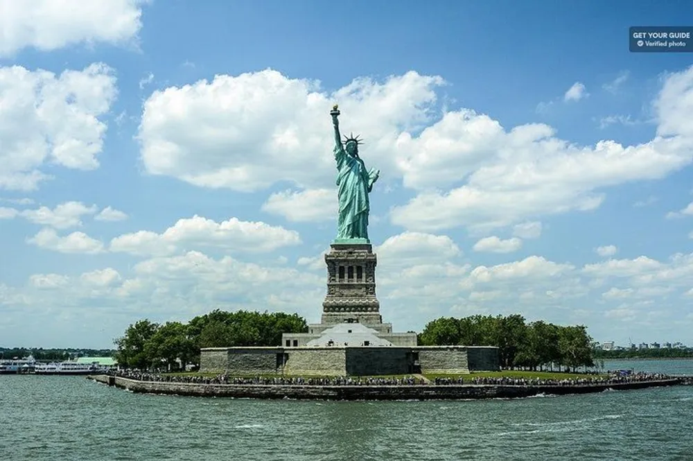 The image shows the Statue of Liberty on a clear day standing tall on Liberty Island with a view of the surrounding water and a partly cloudy sky