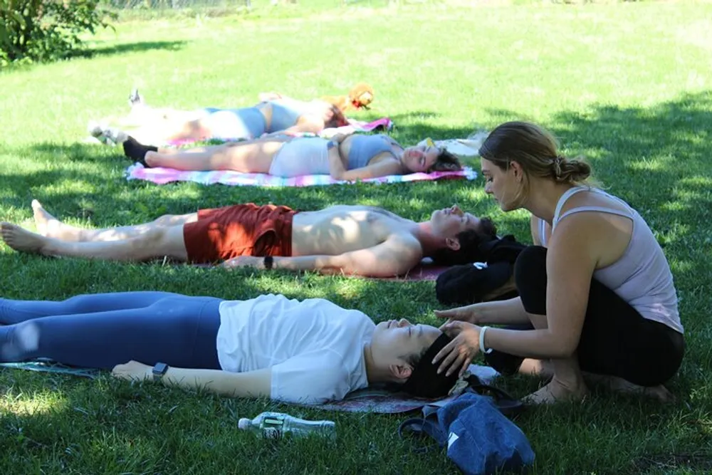 A group of people are lying down on mats in a grassy area while one person appears to be assisting or instructing another person