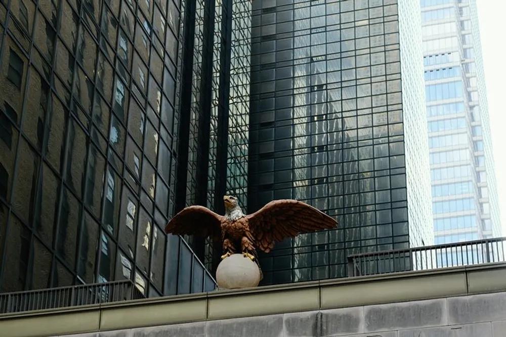 An eagle sculpture with outstretched wings perched on a sphere is set against the backdrop of modern high-rise buildings with reflective glass facades