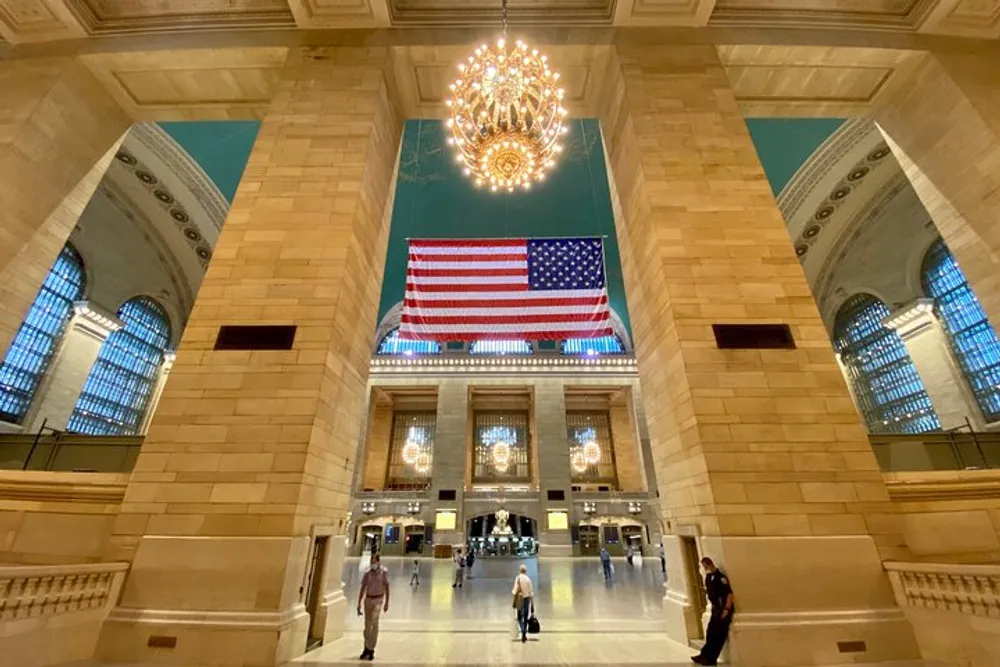 The image shows the spacious and grand interior of a train station with high ceilings a large American flag and a chandelier with a few people walking around