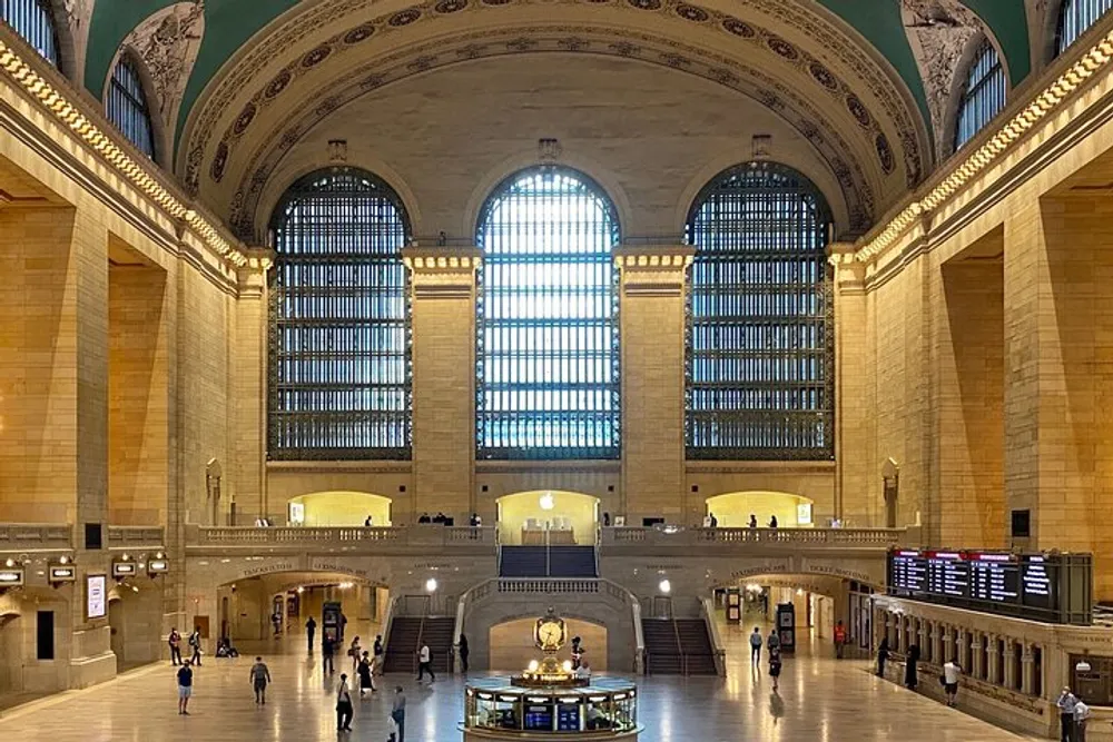 The image shows the interior of a grand historic train station with high ceilings tall windows and a clock in the center above an information booth with several people walking around