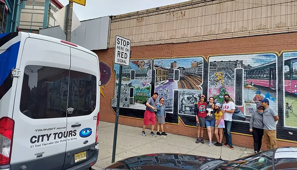 A group of people are posing for a photograph in front of a colorful mural depicting urban scenes with a tour van parked nearby