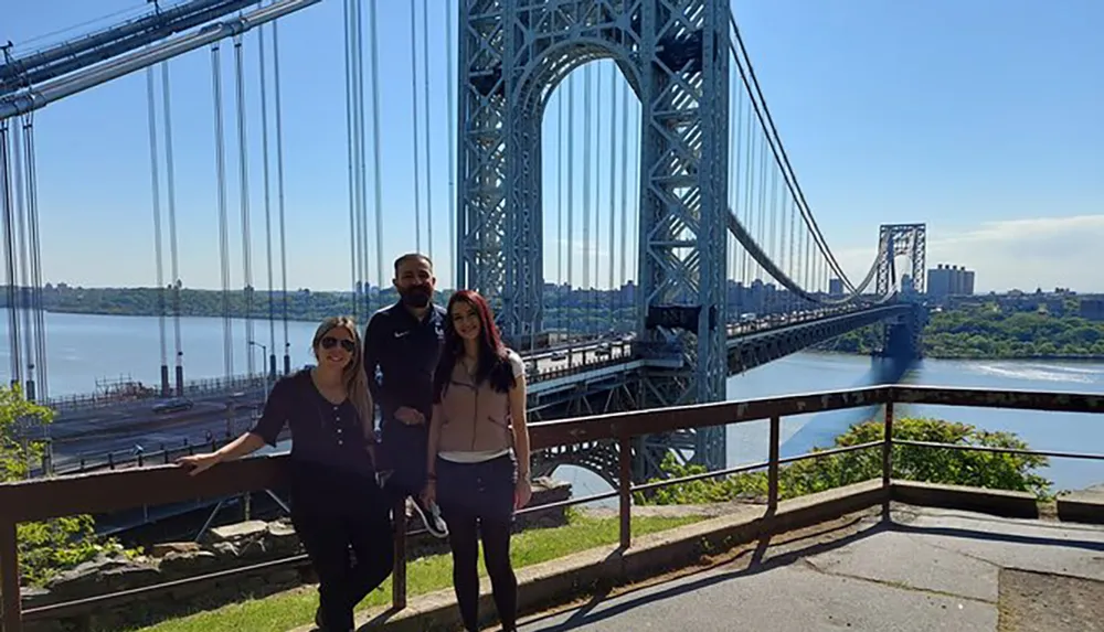 Three people are posing for a photo in front of a striking suspension bridge on a sunny day