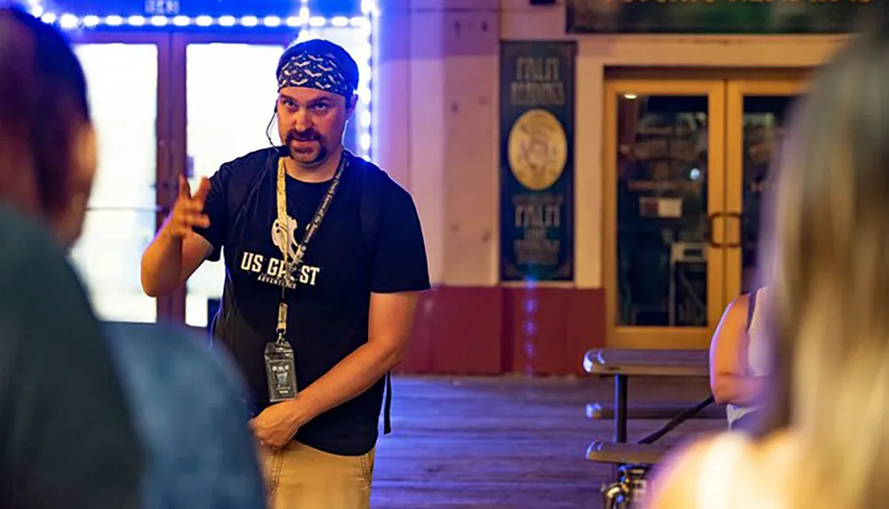 A man wearing a bandana and a T-shirt labeled US GHOST ADVENTURES is gesturing and speaking to an unseen group in what appears to be an evening setting