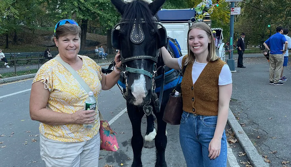 Two smiling women pose beside a horse that is part of a carriage setup in a park-like setting