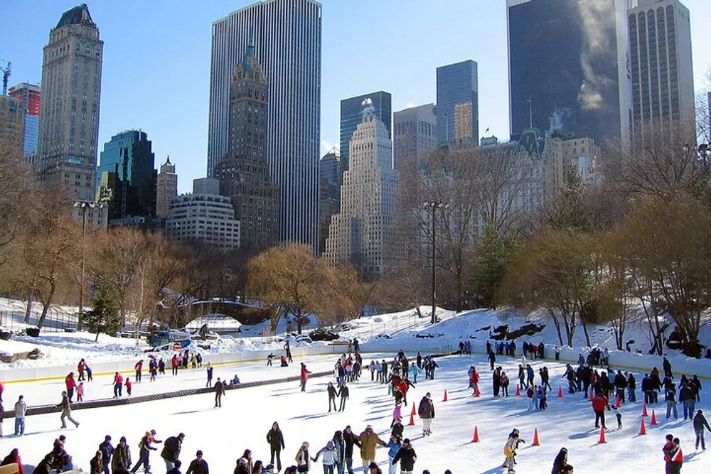 People are ice skating at a rink in a park with tall city buildings in the background on a bright winter day
