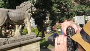 A photographer is capturing a scene featuring a lion statue, with various other classical statues and a garden in the background.