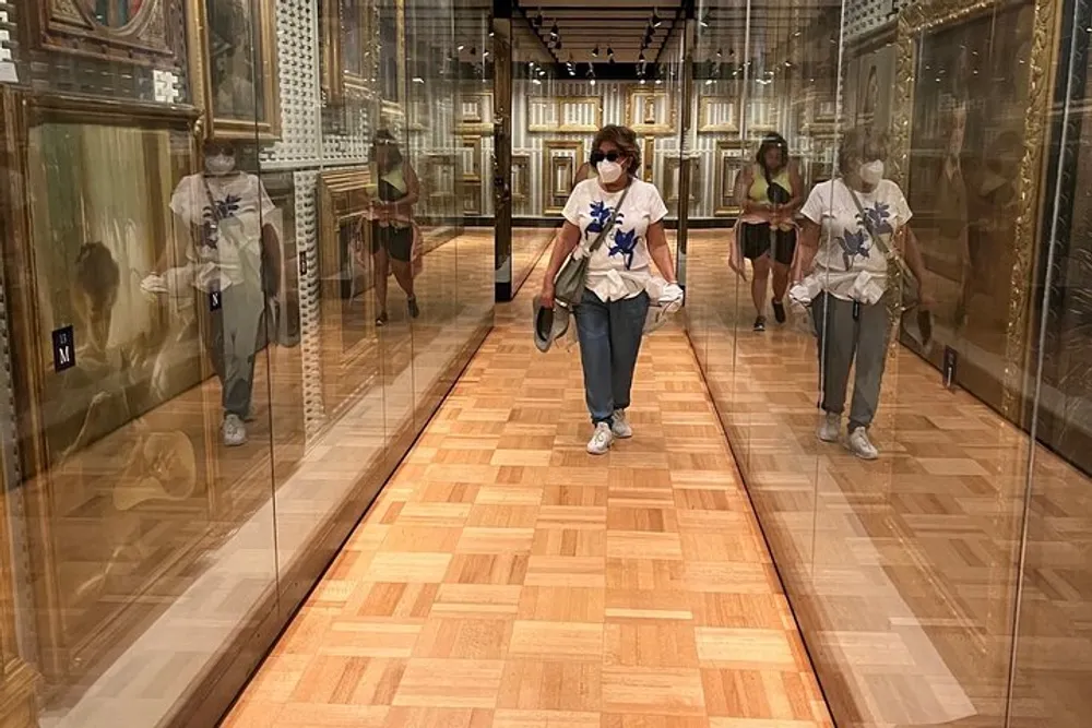 A person wearing a mask is walking through a gallery with multiple reflections visible in the glass display cases on either side
