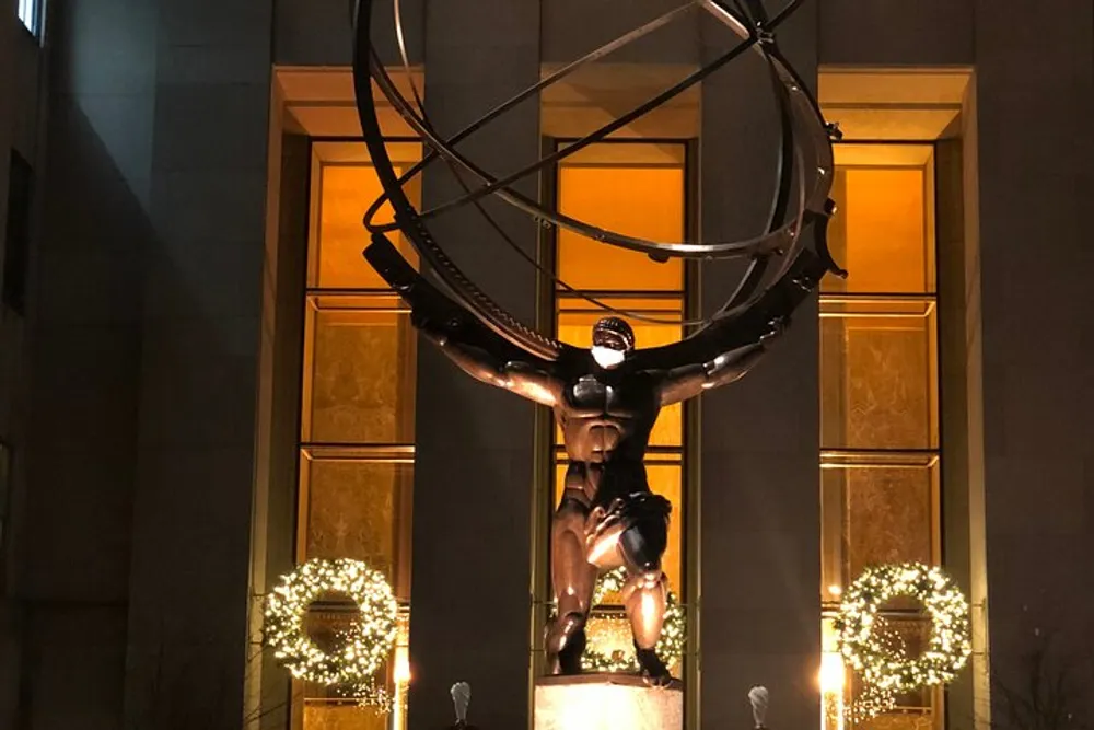 The image shows a large bronze sculpture of Atlas holding celestial spheres on his back set against a building with illuminated windows and festive wreaths