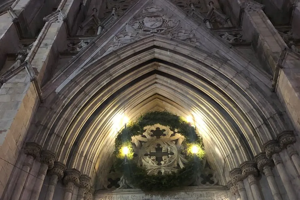 The image shows an intricately carved archway of a Gothic-style stone building adorned with a green wreath and lit by ambient lighting