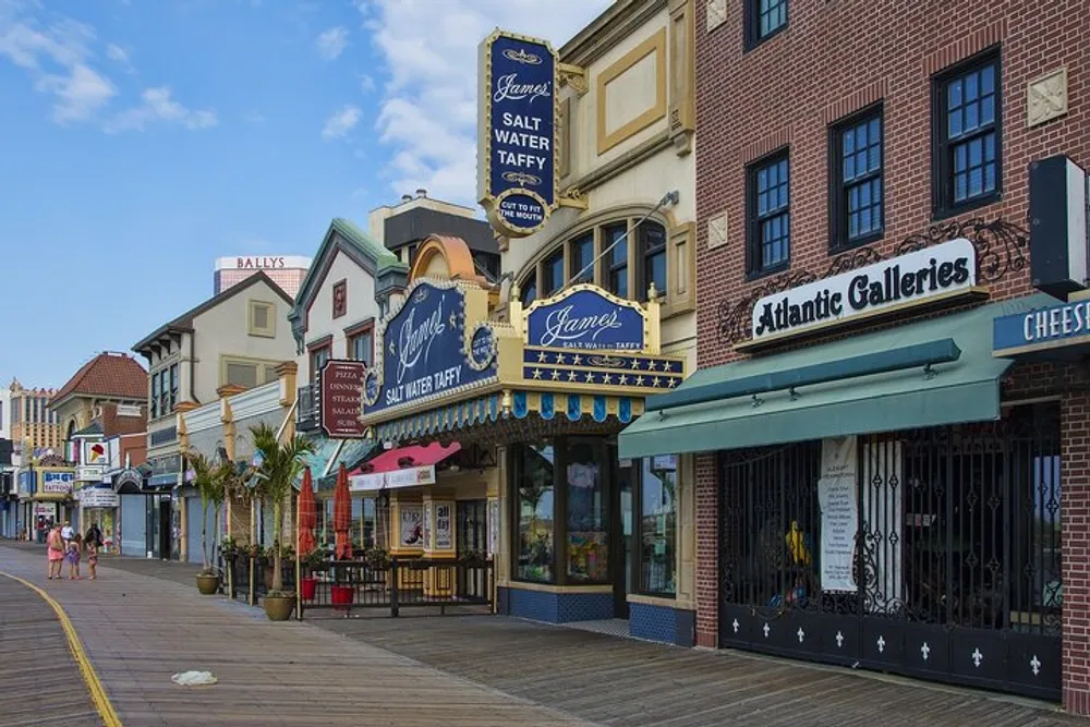 The image shows a sunny boardwalk lined with colorful storefronts including one advertising salt water taffy with a few people strolling by