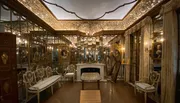 The image shows an ornate room with gilded mirrors and intricate decorations, featuring classical portraits, a chandelier, and richly designed furniture, evoking a sense of historical opulence.