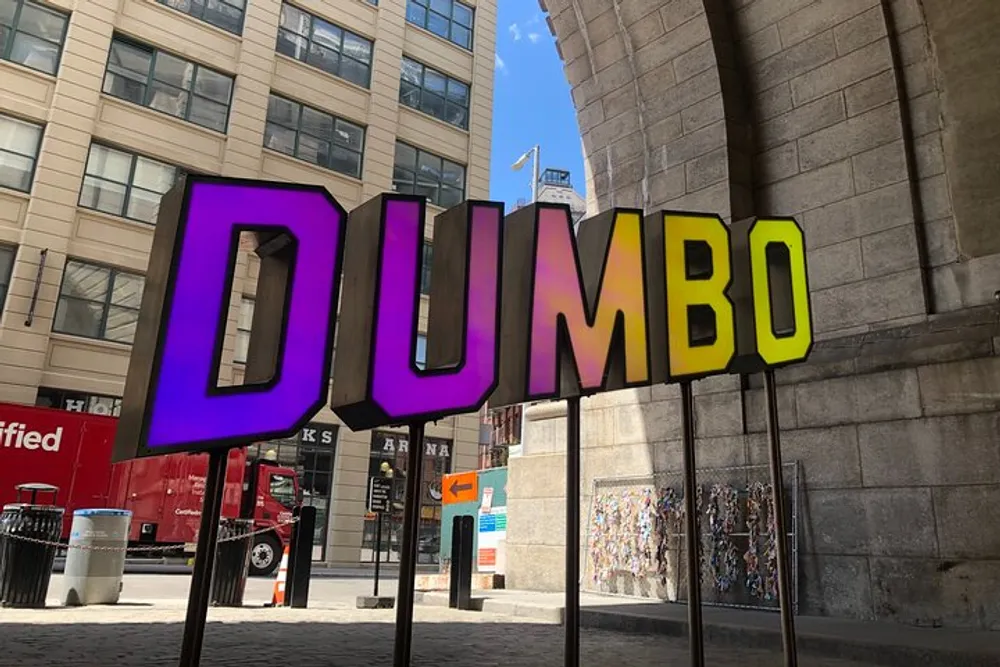 The image shows a colorful large-scale sign spelling DUMBO on display in an urban setting with buildings and a clear sky in the background