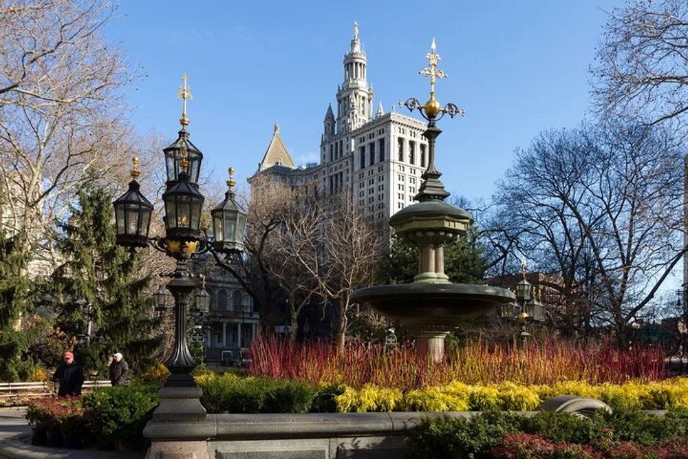 An ornate fountain and vintage street lamps stand amidst colorful plant life in an urban park with a tall historic building in the background