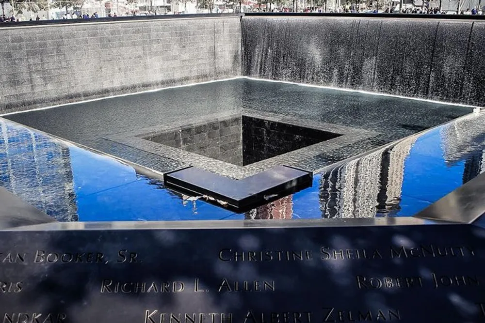 The image depicts a large square memorial pool with water cascading down its sides surrounded by panels with inscribed names as part of a somber commemorative site