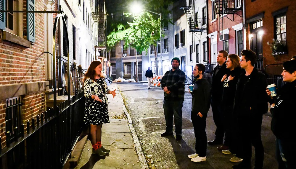 A group of people are attentively listening to a woman who appears to be giving a tour at night on an urban street