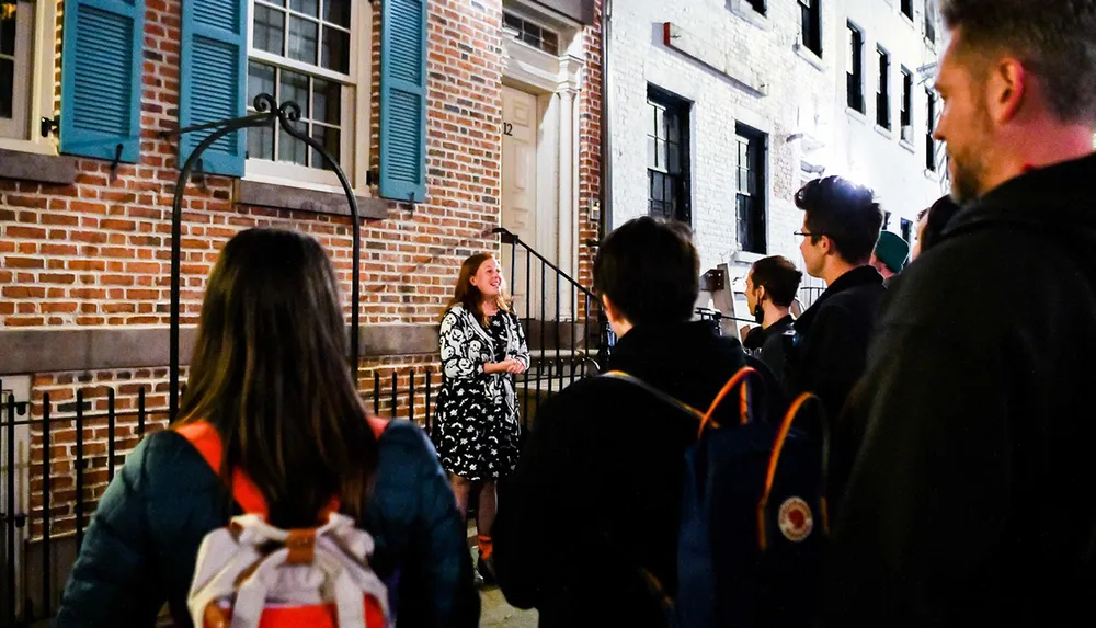 A woman appears to be giving a tour or presentation to a group of attentive onlookers at night outside a building with brick walls and blue shutters