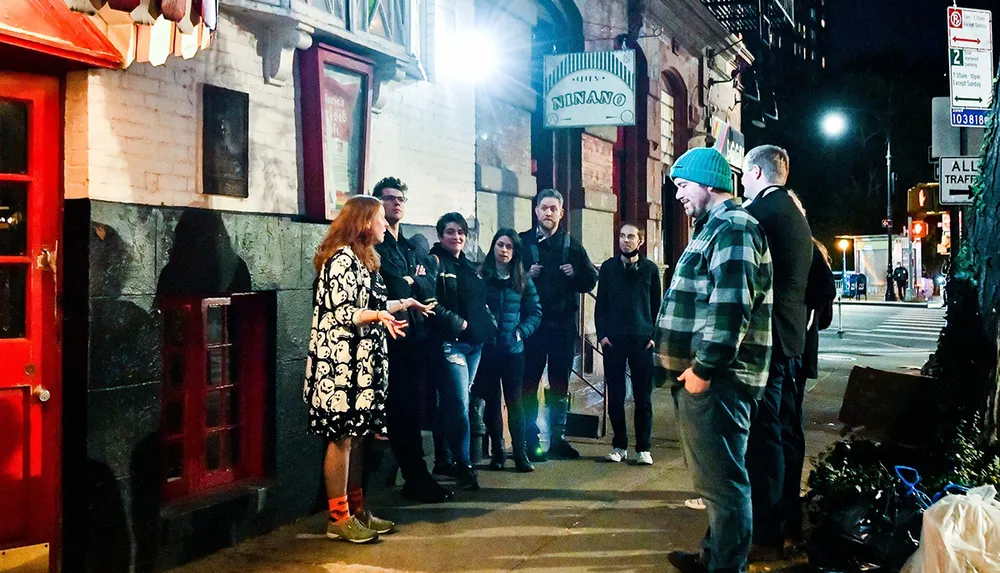 A group of people is standing on a city sidewalk at night outside a building with a sign reading NINANO engaged in a conversation