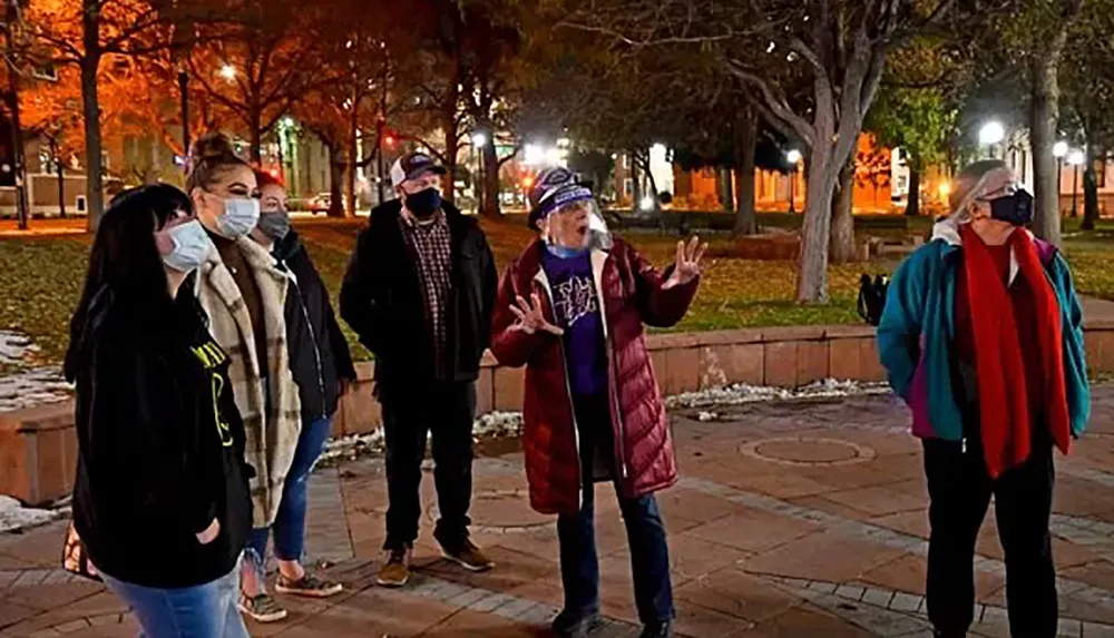 A group of people some wearing masks are engaged in a conversation or tour outdoors in an urban park at night