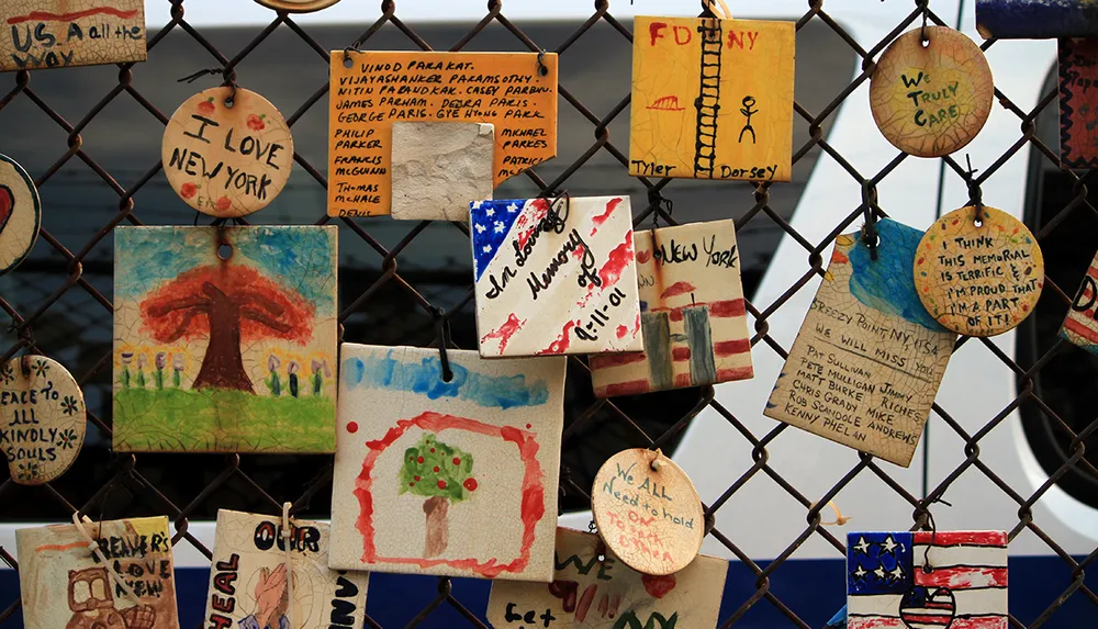 The image shows a collection of handcrafted tiles and circular plaques with various messages and artwork including tributes to New York displayed on a chain-link fence