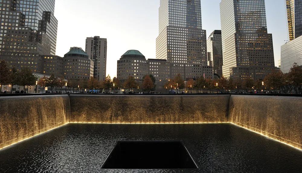 The image shows one of the 911 Memorial pools at the World Trade Center site in New York City with surrounding skyscrapers bathed in the warm glow of dusk