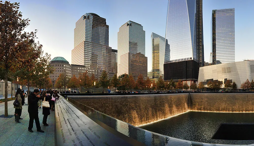 The image captures visitors at the 911 Memorial reflecting pools with the names of the victims inscribed on the bronze parapets set against the backdrop of the new World Trade Center buildings in Lower Manhattan at sunset