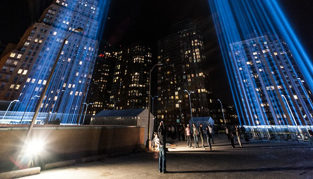 The image shows a nighttime scene with people gathering around beams of light shooting into the sky amidst urban high-rise buildings