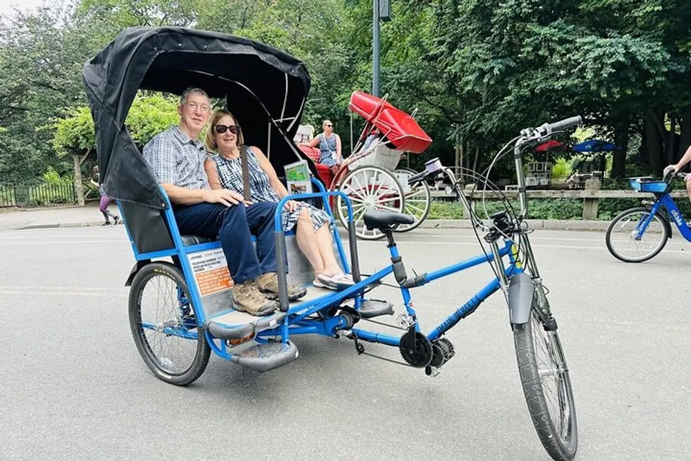 Two people are enjoying a ride on a blue pedicab through a park while another individual bikes in the background