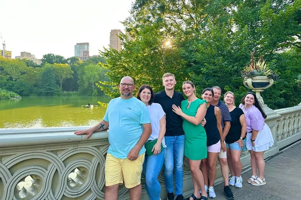 A group of seven people are smiling and posing for a photo on a decorative bridge with a lush green park and a pond in the background