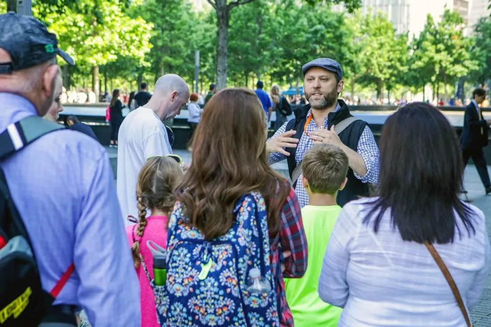 A man in a cap is speaking to a group of onlookers in a sunlit outdoor setting with trees in the background