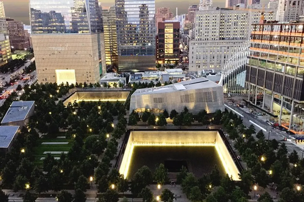 The image shows a high-angle view of the 911 Memorial in New York City at dusk with the illuminated twin reflecting pools surrounded by trees and bordered by modern buildings