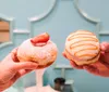 Two hands are holding up delicious-looking doughnuts one topped with powdered sugar and a strawberry slice and the other with orange icing and white drizzle against a teal background