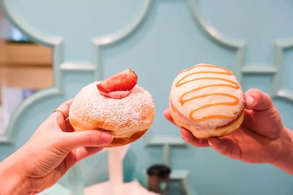Two hands are holding up delicious-looking doughnuts one topped with powdered sugar and a strawberry slice and the other with orange icing and white drizzle against a teal background