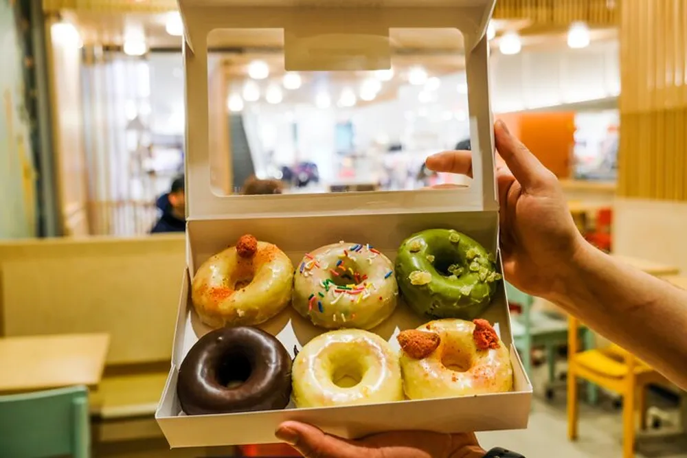 A person is holding an open box of six assorted doughnuts inside a brightly lit cafe
