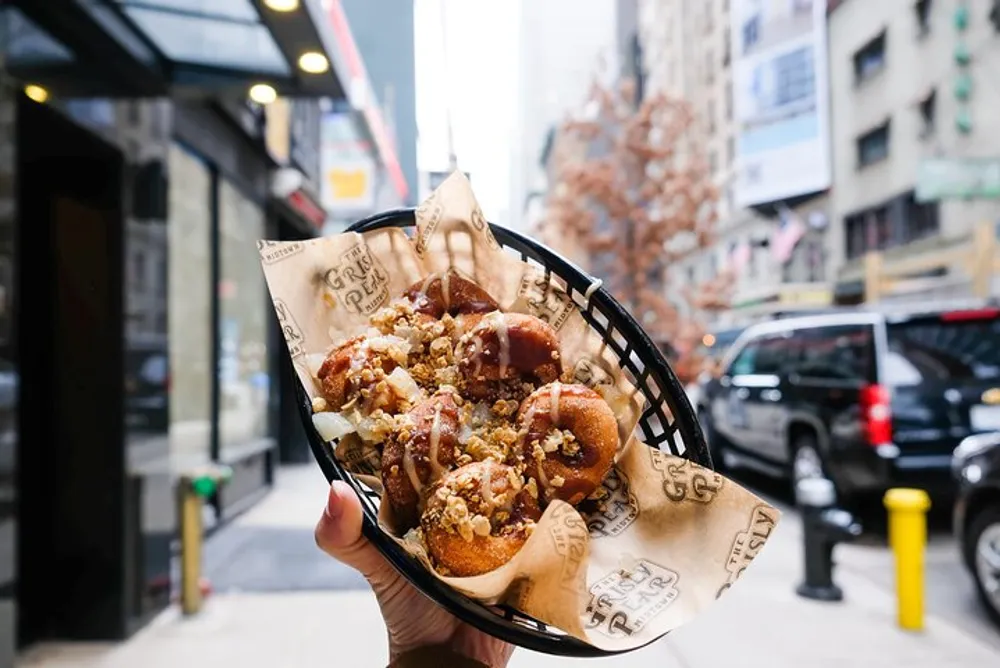 A person is holding a basket of pretzel bites drizzled with sauce and sprinkled with nuts against an urban street backdrop