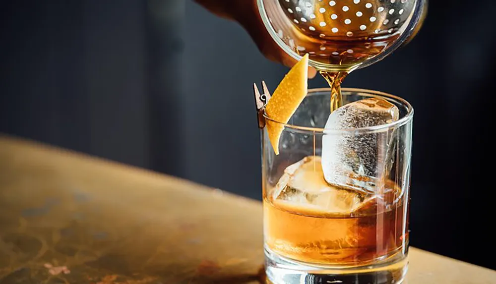 A cocktail is being poured into a glass with a large ice cube and garnished with a twist of citrus peel