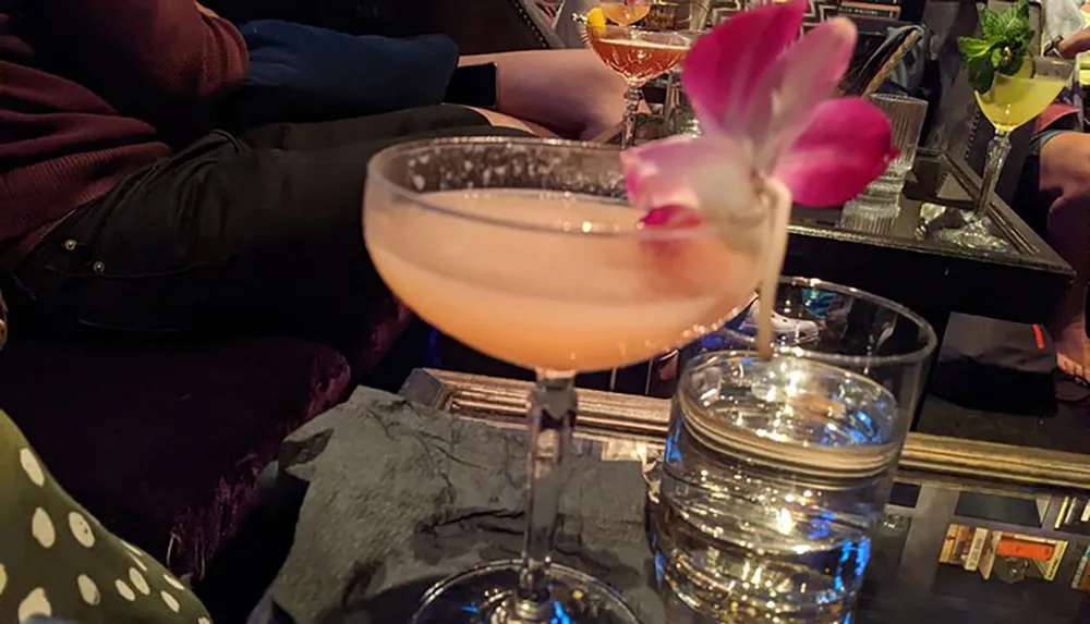 A pink cocktail garnished with a flower is in focus with people and other drinks slightly blurred in the background at a bar setting