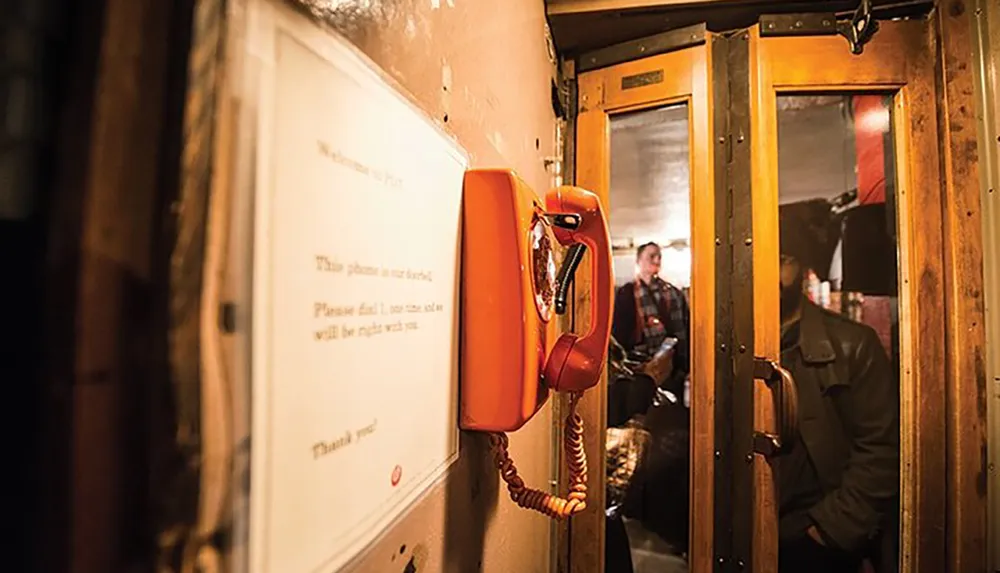 The image shows a vintage orange rotary phone inside a wooden phone booth with a sign indicating the phone is a direct line while people sit in the background possibly in a restaurant or bar setting