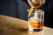 A cocktail is being poured into a glass containing a large ice sphere, garnished with a twist of citrus peel secured by a mini clothespin.