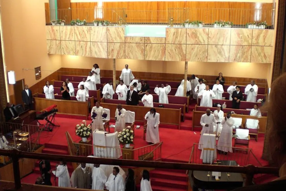 The image shows a congregation most wearing white robes gathered inside a church with wooden pews and a pulpit indicating a religious ceremony or service in progress